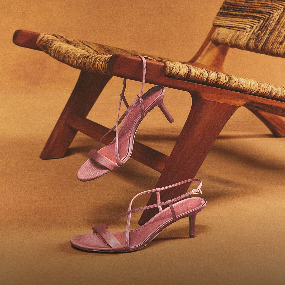Pink Ginny strappy sandals with a low heel displayed on and beside a woven wooden chair.