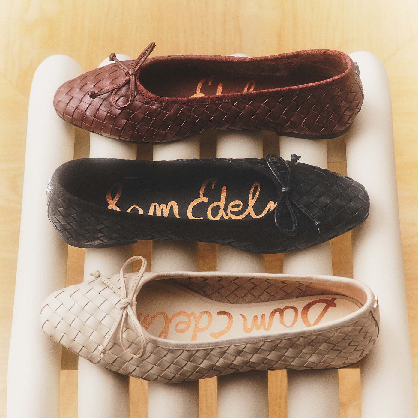 Three Sam Edelman Ruthie woven ballet flats with bow details in brown, black, and ivory, arranged on a wooden slatted surface.